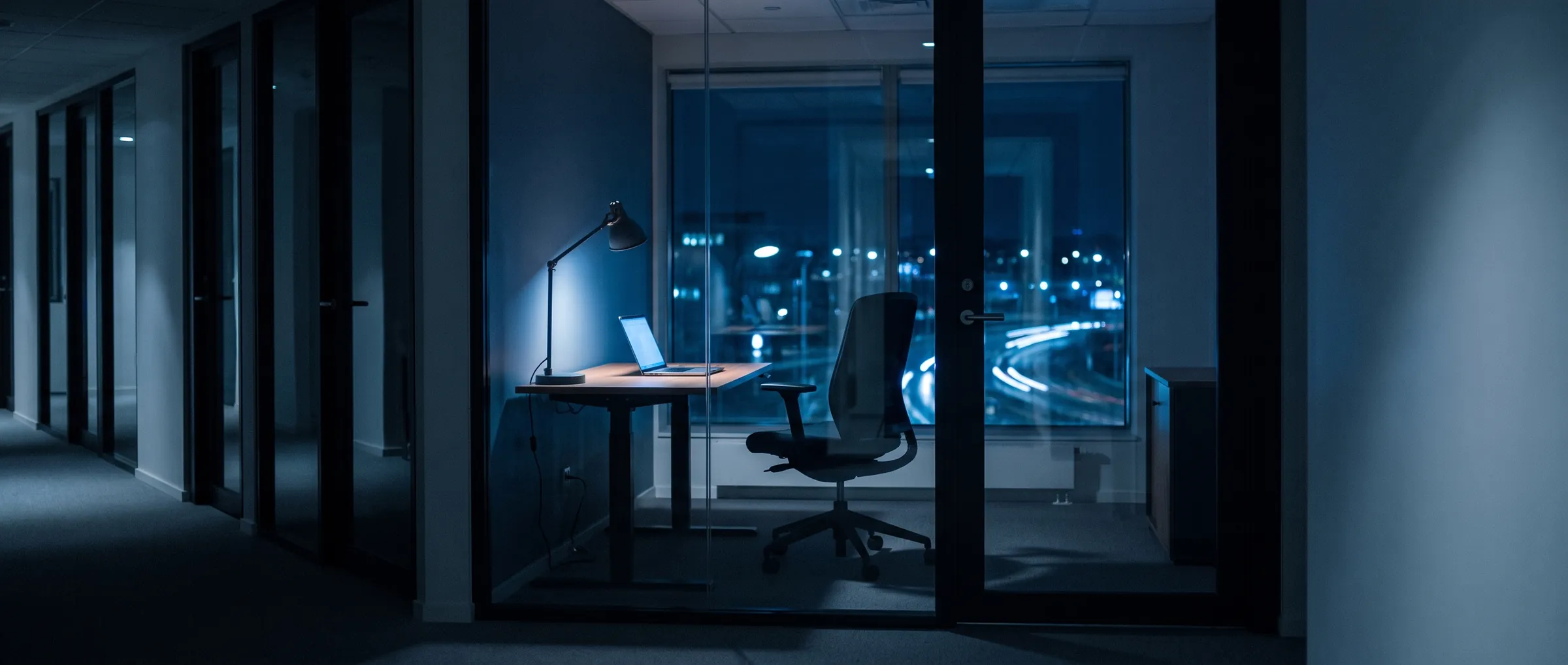 Sala de escritório à noite, com uma luminária acesa sobre a mesa e a cidade ao fundo pela janela